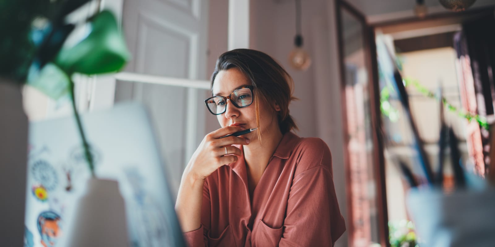 A woman at her laptop in a sunlit room, a pen held against her mouth, pausing before she answers